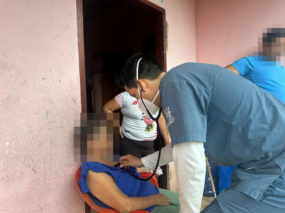 El Dr. Raúl Muñoz, de CrabCare, ausculta a una paciente adulta mayor sentada en la puerta de su casa, con su familia al lado