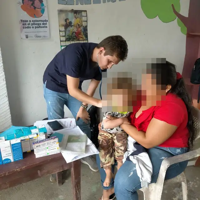 Un médico de CrabCare ausculta a un niño pequeño en brazos de su madre, durante una atención a domicilio